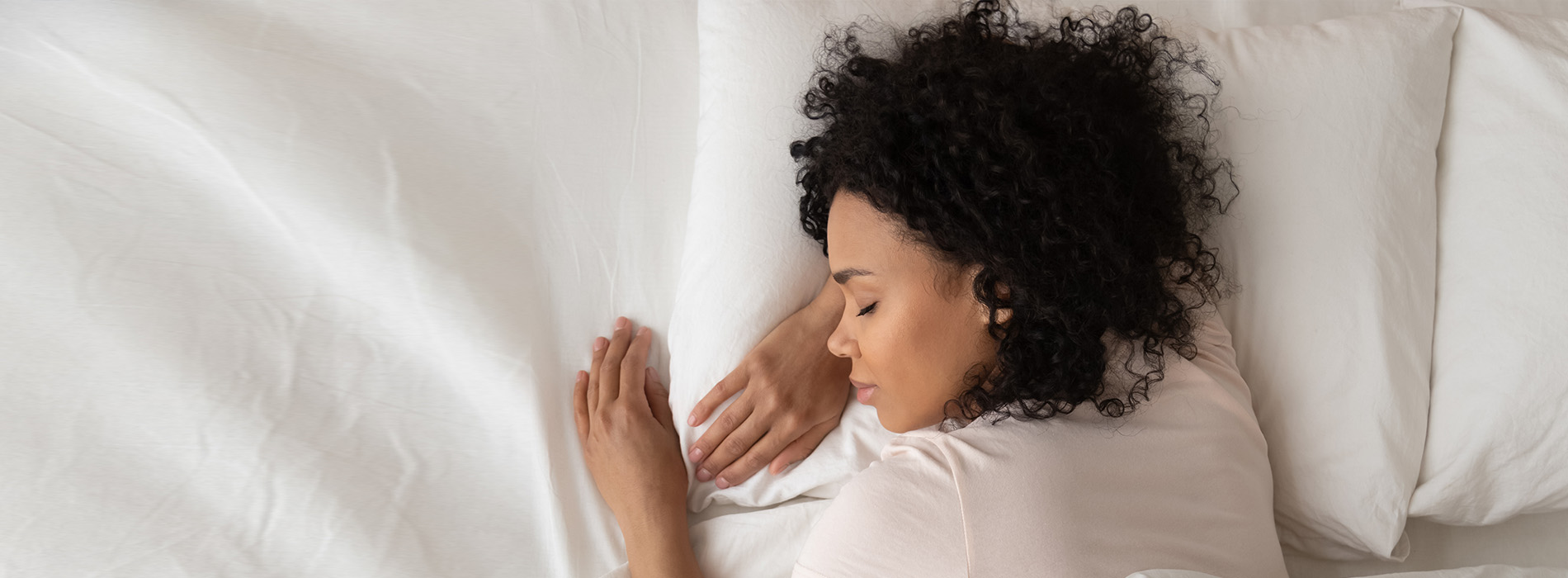 A photograph of a person lying in bed, with their head resting on a pillow and hands clasped together. The individual appears to be in a state of relaxation or possibly deep thought.