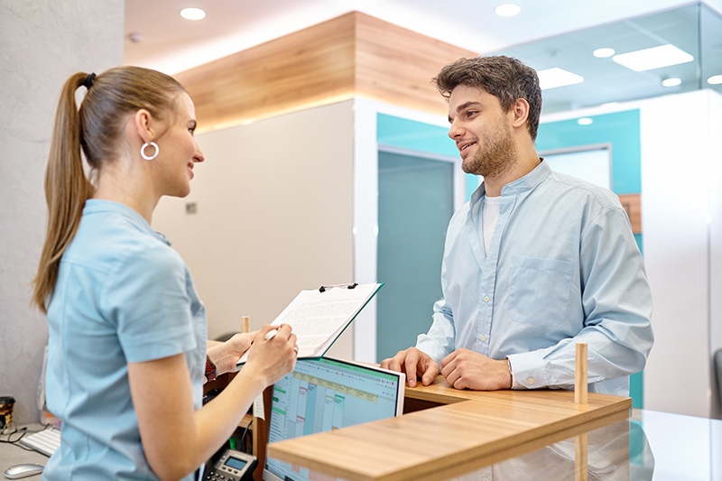 A man and a woman in a professional setting, with the man standing behind a desk and the woman seated on the other side, both engaged in conversation.